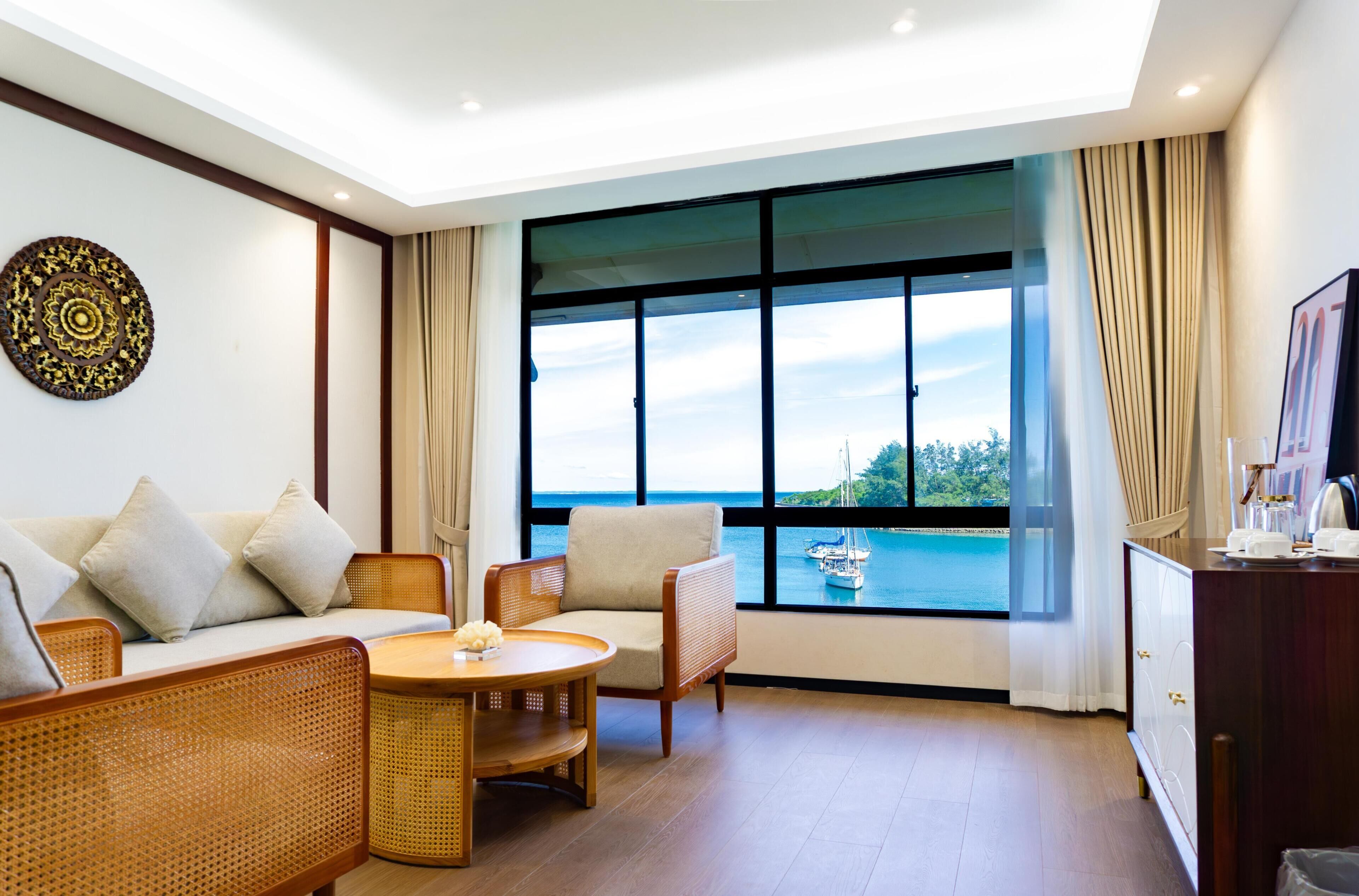 A room with a sofa, two chairs, a small table, a decorative wall piece, and a large window with a view of the sea and boats.