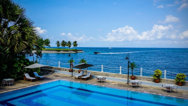 A swimming pool with lounge chairs and tables, overlooking a beach with palm trees and a view of the ocean.
