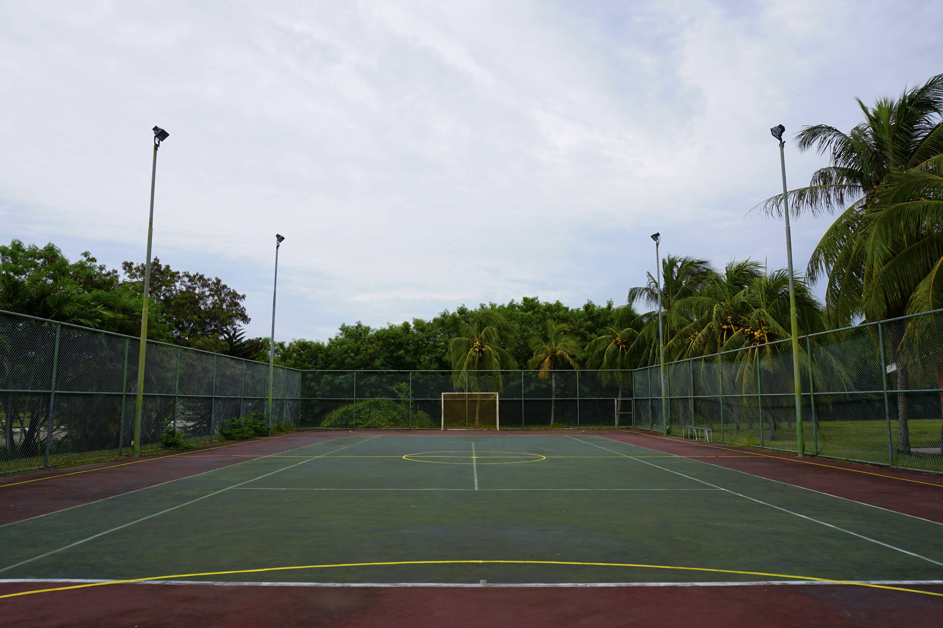 An outdoor tennis court with a net, surrounded by a fence and palm trees.