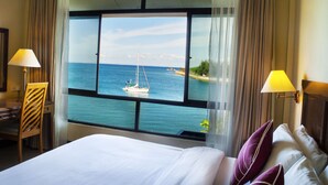 A hotel room with a large window offering a view of the sea and a sailboat. There is a bed with pillows and a desk with a lamp.
