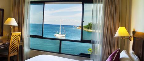 A hotel room with a large window offering a view of the sea and a sailboat. There is a bed with pillows and a desk with a lamp.
