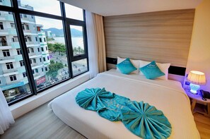 A hotel room with a large window, a bed with teal decorative pillows, and a bedside table with a lamp. - The MCR Luxury Nha Trang Hotel (Nha Trang)