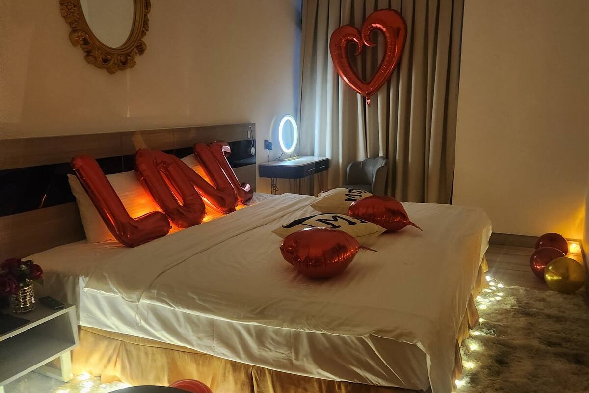 Habitación de hotel con cama, mesita de noche, espejo, escritorio con lámpara y un globo con forma de corazón.