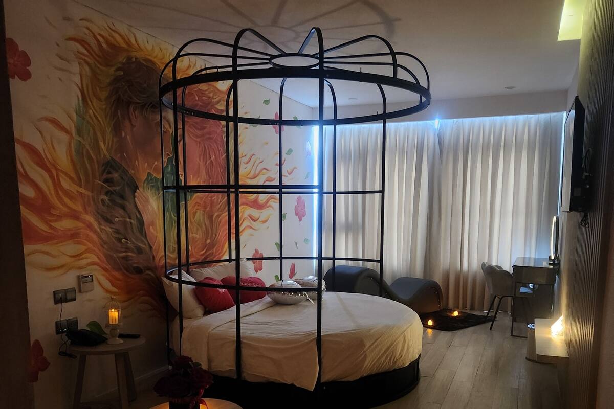 Una habitación de hotel moderna con una cama redonda, un mural decorativo, una mesita con un jarrón de flores y un escritorio con una lámpara.