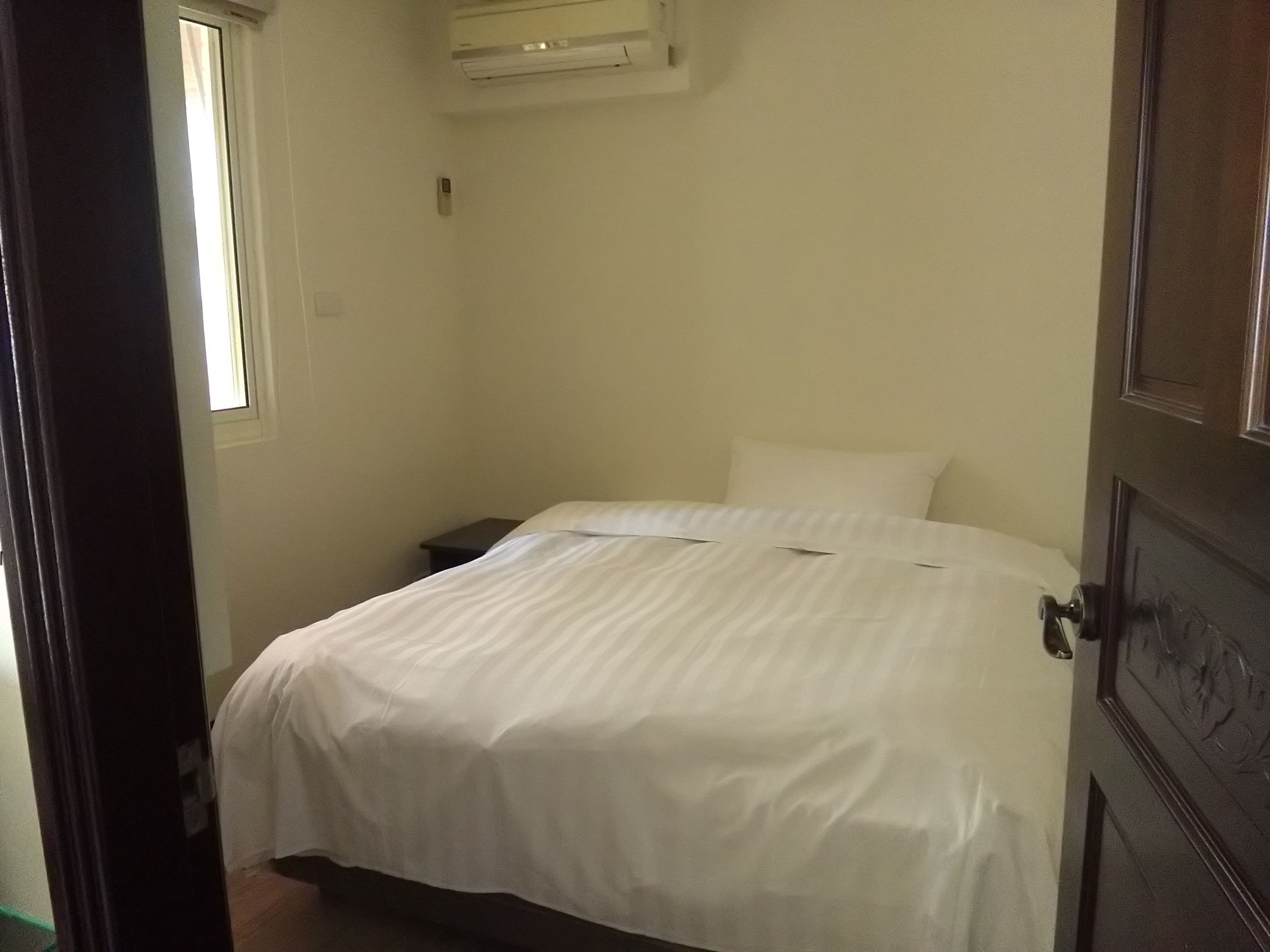 Una cama pequeña y bien arreglada en una habitación con ventana, una mesita de noche y un equipo de aire acondicionado en la pared.