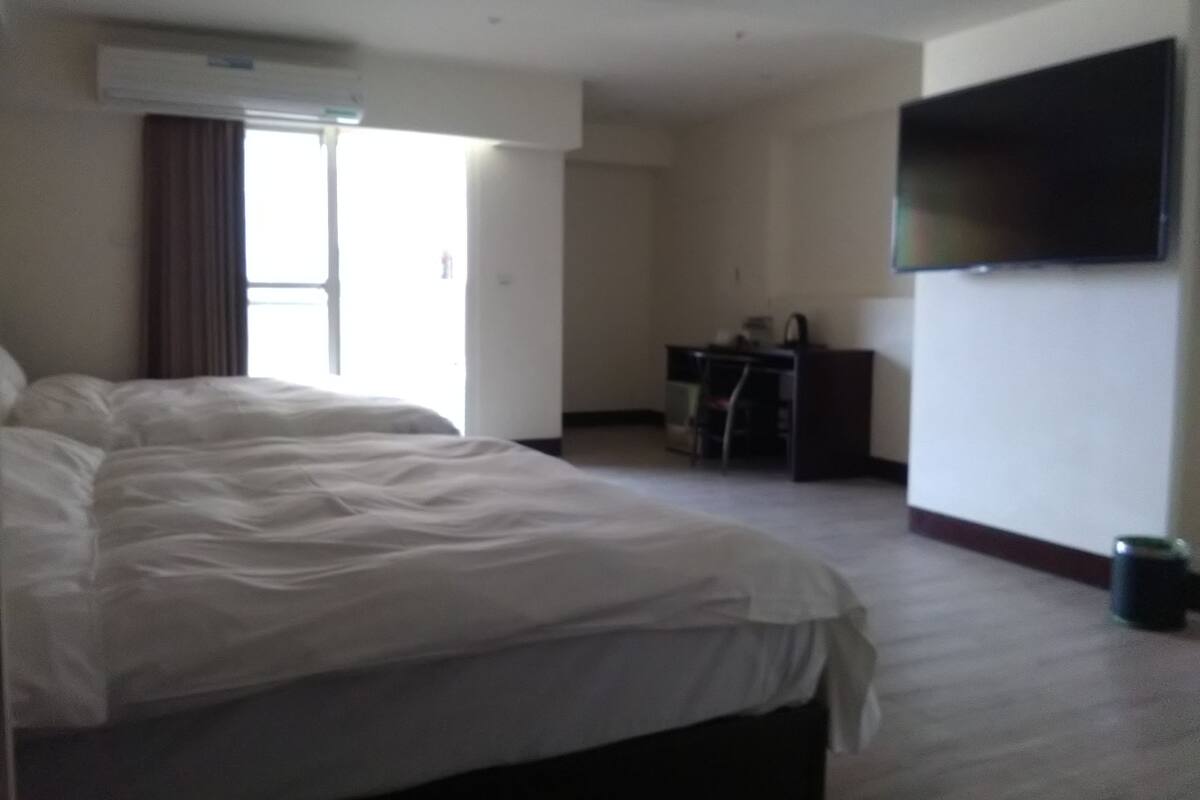 A hotel room with a large bed, a desk, a chair, a television mounted on the wall, and an air conditioning unit.