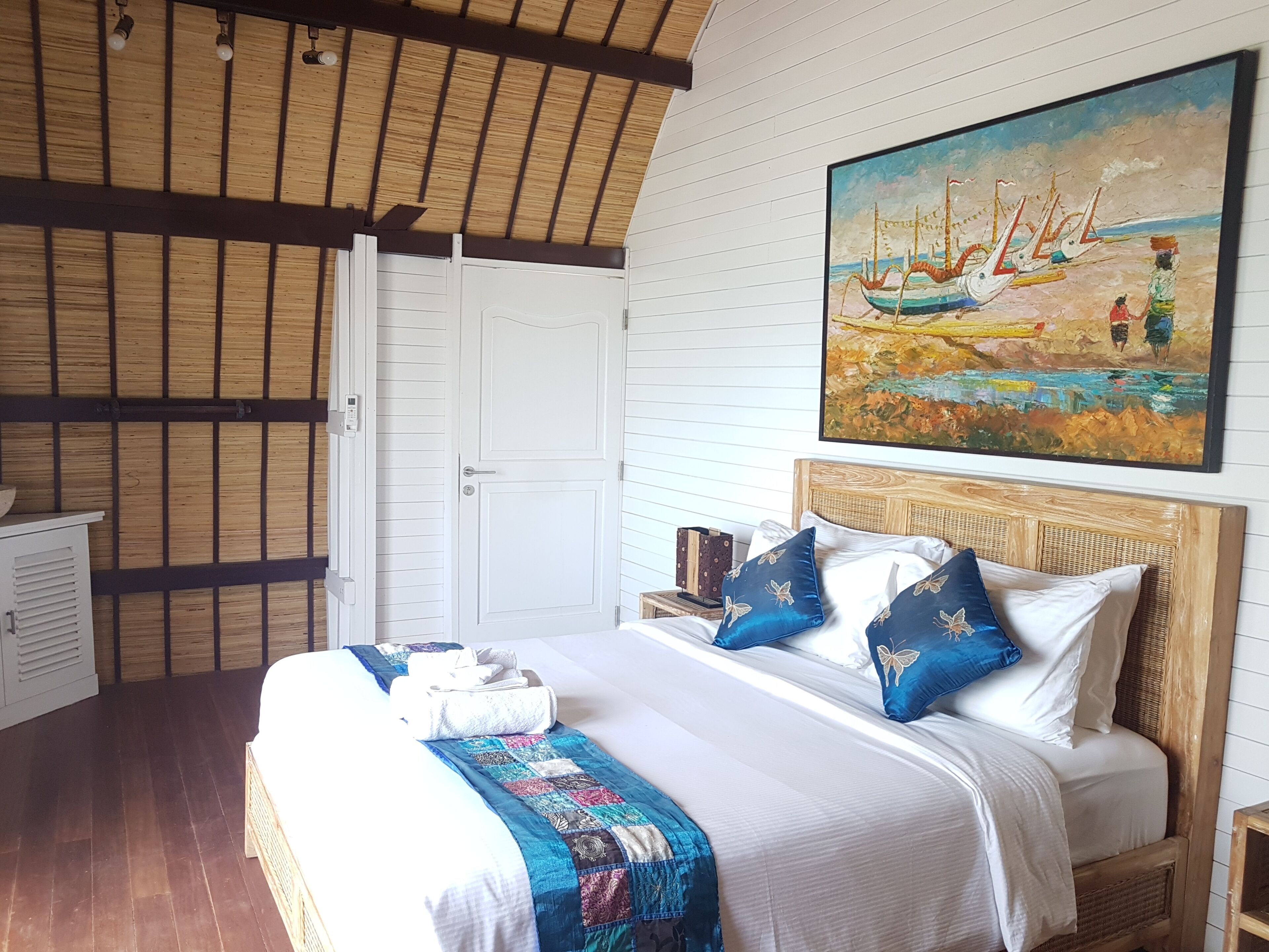 A bedroom with a wooden ceiling, a bed with white bedding and blue pillows, a painting of boats on the wall, and a white wardrobe.