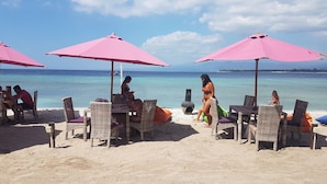 Una spiaggia con sedie e tavoli in vimini sotto ombrelloni rosa, affacciata su un mare cristallino.