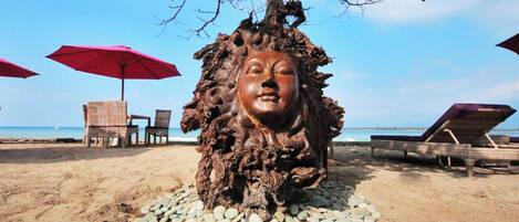 A beach scene with a wooden sculpture of a lion's head surrounded by pebbles, two red umbrellas, and beach chairs.