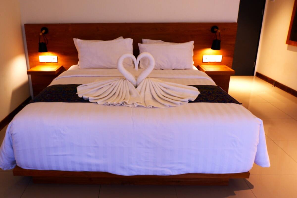 A neatly made bed with a heart-shaped towel arrangement, wooden headboard, and bedside lamps.