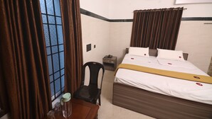 A hotel room with a bed, a chair, a small table with a pitcher, a nightstand, and an air conditioning unit. - GM Residency (Chennai)