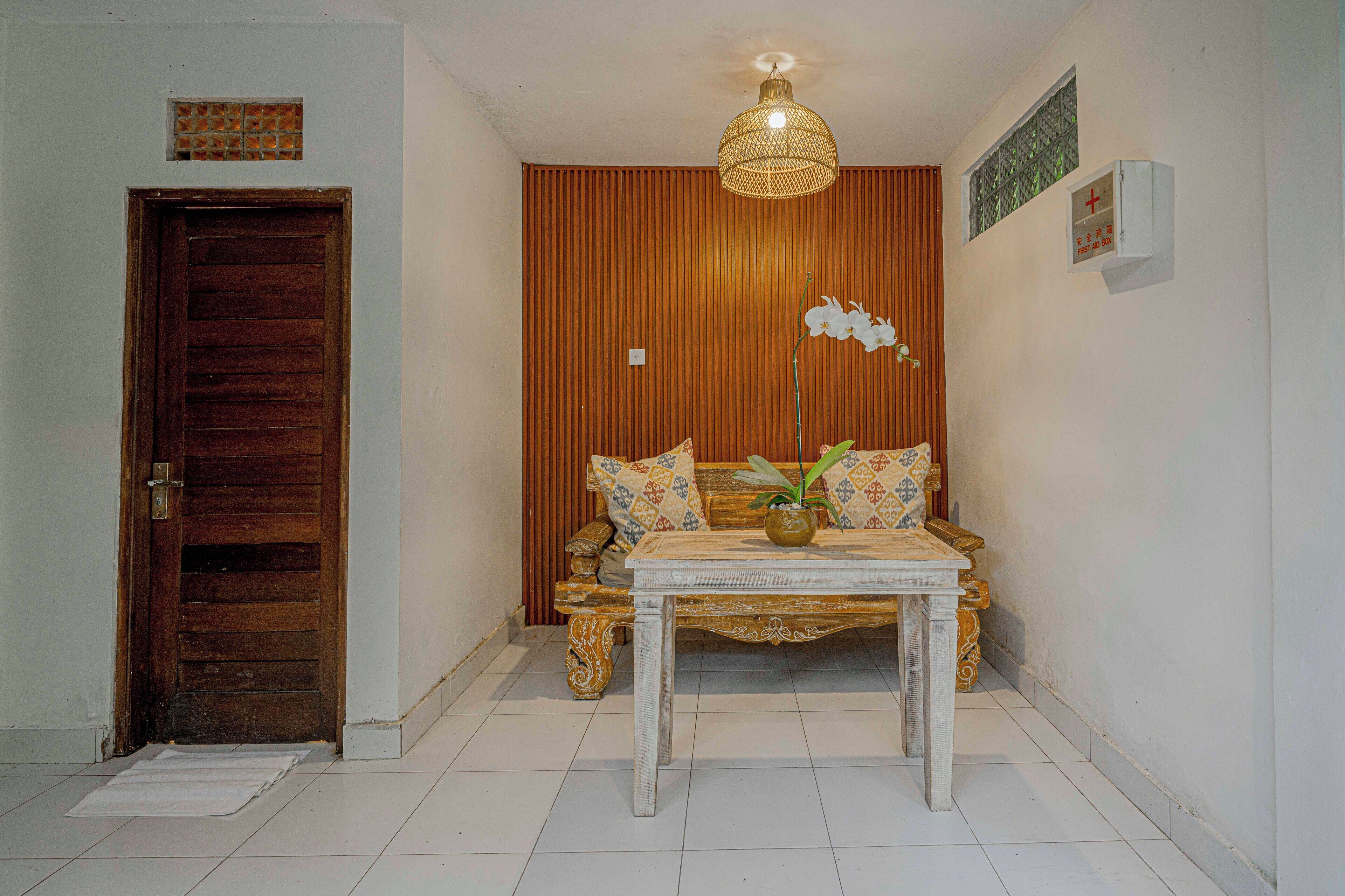 A room with a wooden door, a sofa with patterned cushions, a table with a vase, and a hanging light fixture.