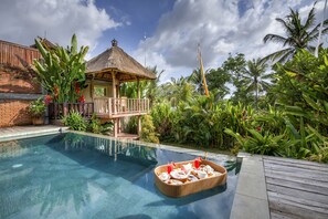 A pool with a thatched-roof hut, lush greenery, and a wooden deck. - Villa Bedauh Ubud (Ubud)