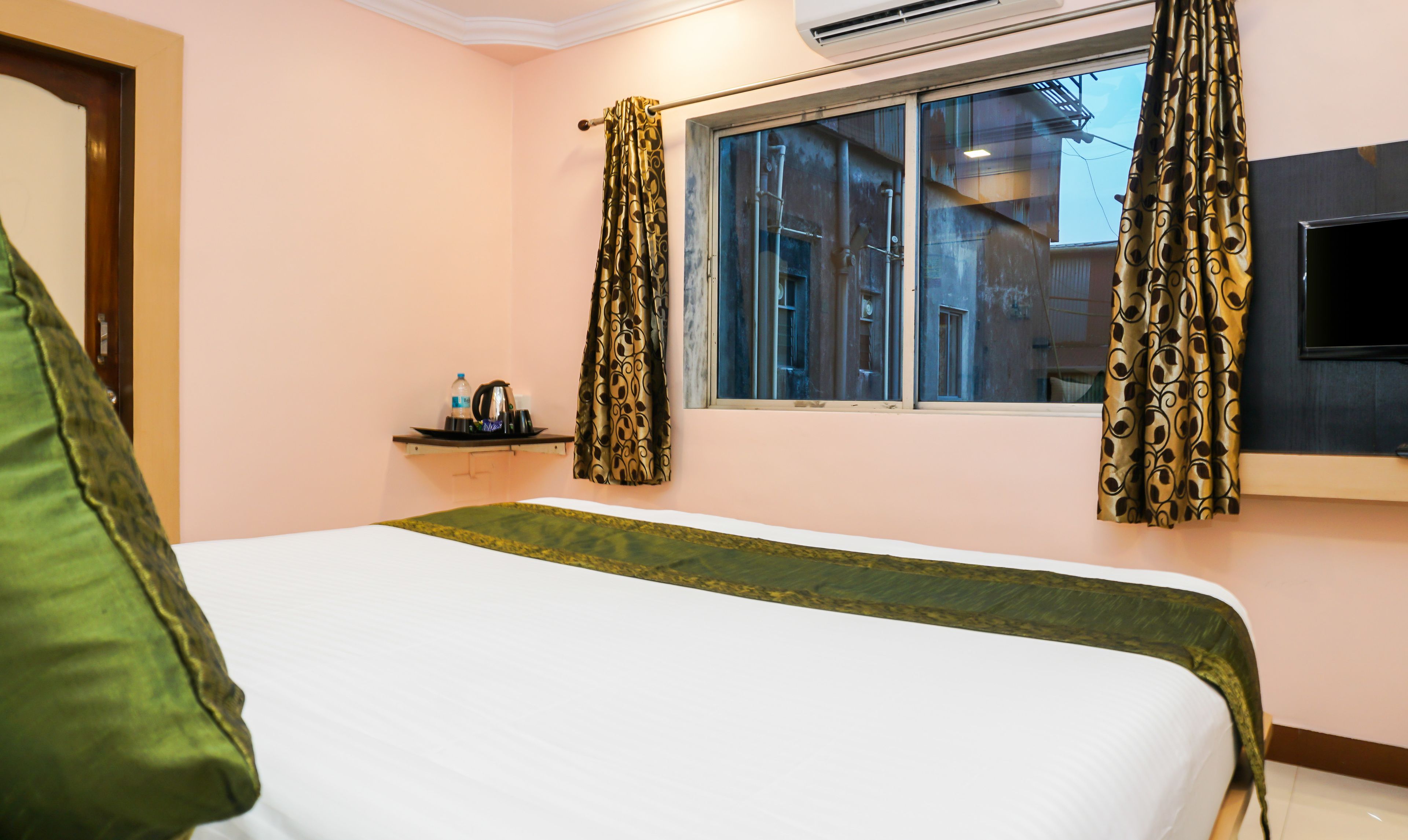 Habitación de hotel con cama, una mesita con una bandeja de objetos, una ventana con cortinas y un televisor de pantalla plana montado en la pared.