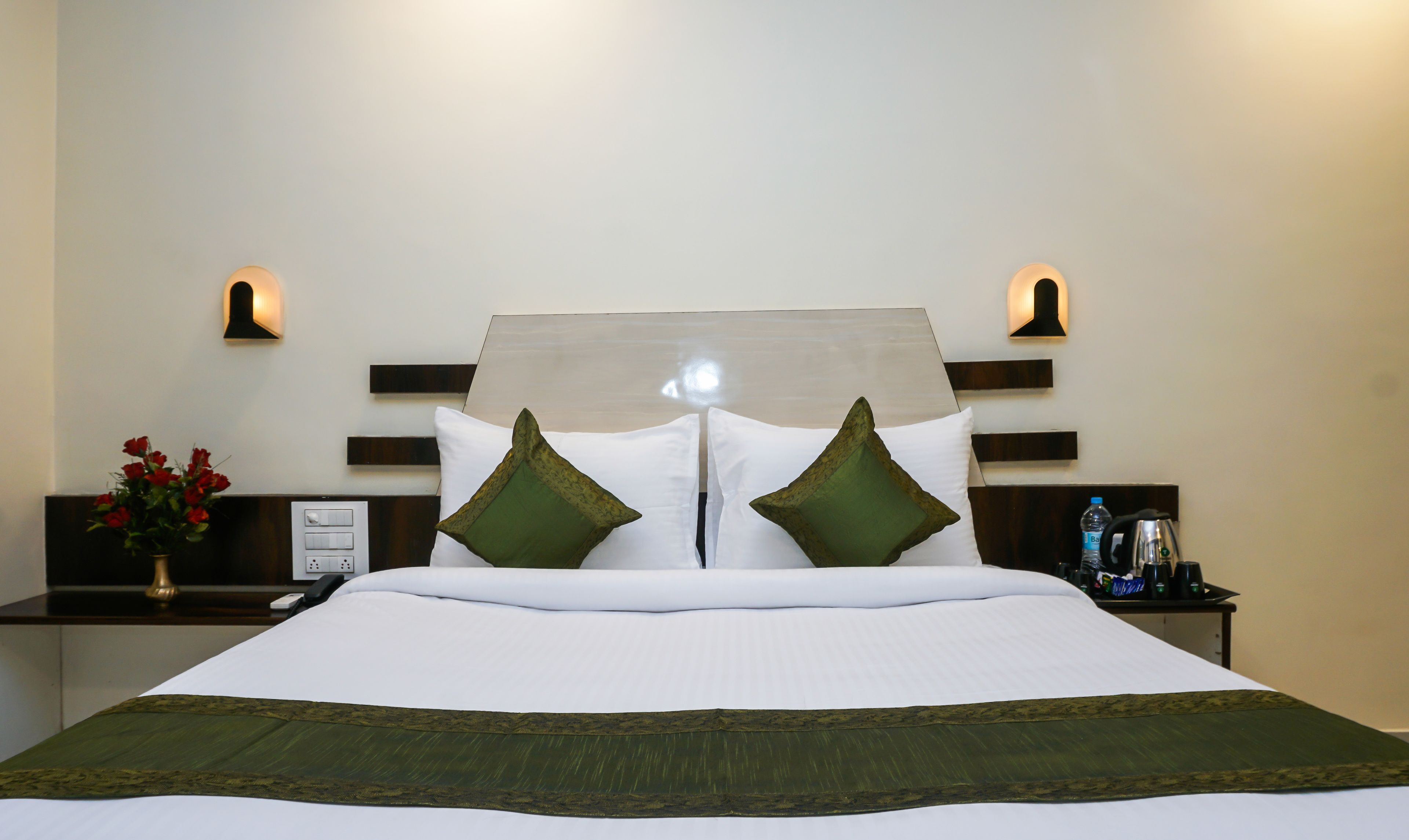 A neatly made bed with white bedding and green decorative pillows, a bedside table with a water bottle and a coffee maker, and wall-mounted light fixtures.