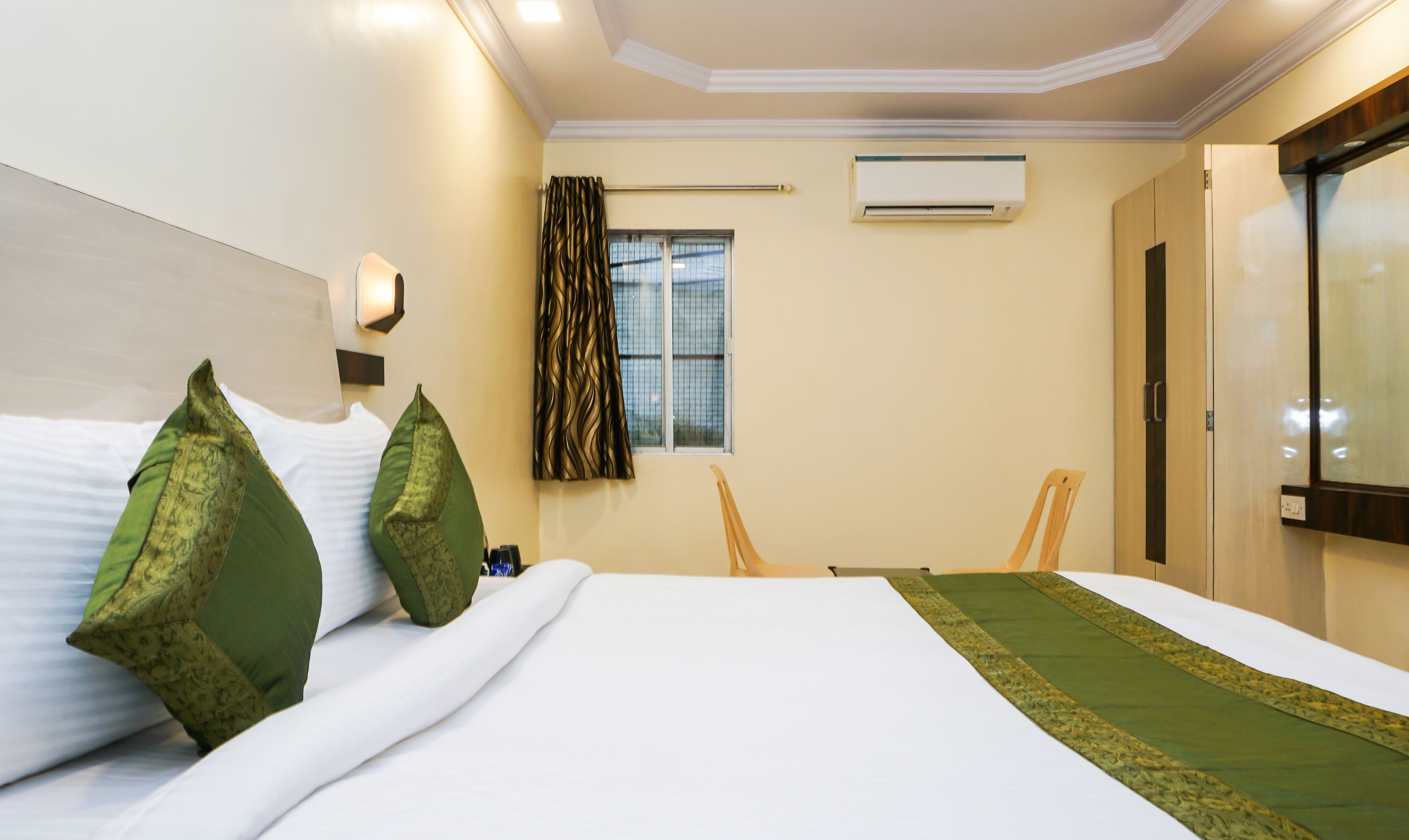 A hotel room with a bed, two chairs, a window with curtains, an air conditioning unit, and a door.