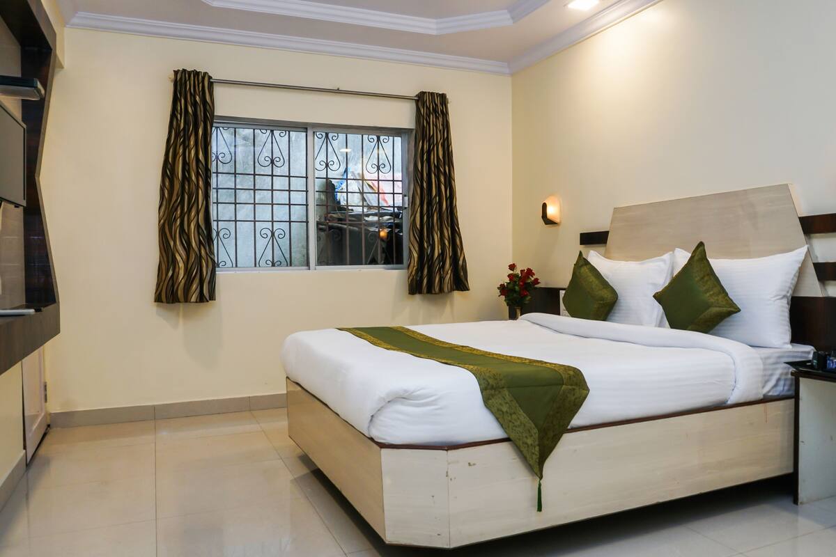 A neatly made bed with white linens and a green throw, a bedside table with a water bottle and glasses, and a window with curtains.