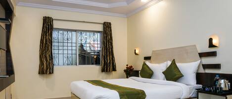 A neatly made bed with white linens and a green throw, a bedside table with a water bottle and glasses, and a window with curtains.
