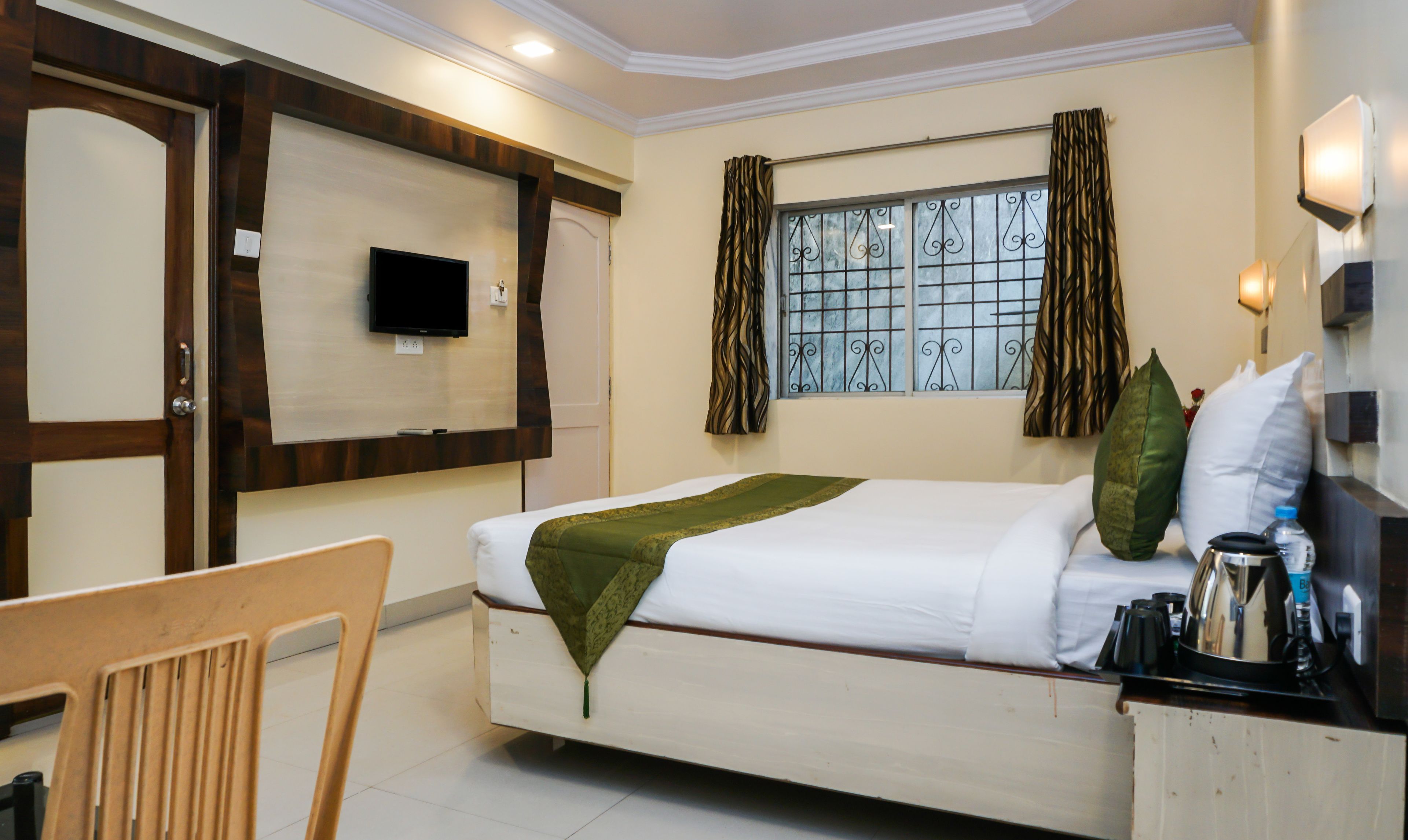 A hotel room with a bed, a television mounted on the wall, a window with curtains, a chair, and a bedside table with a kettle.