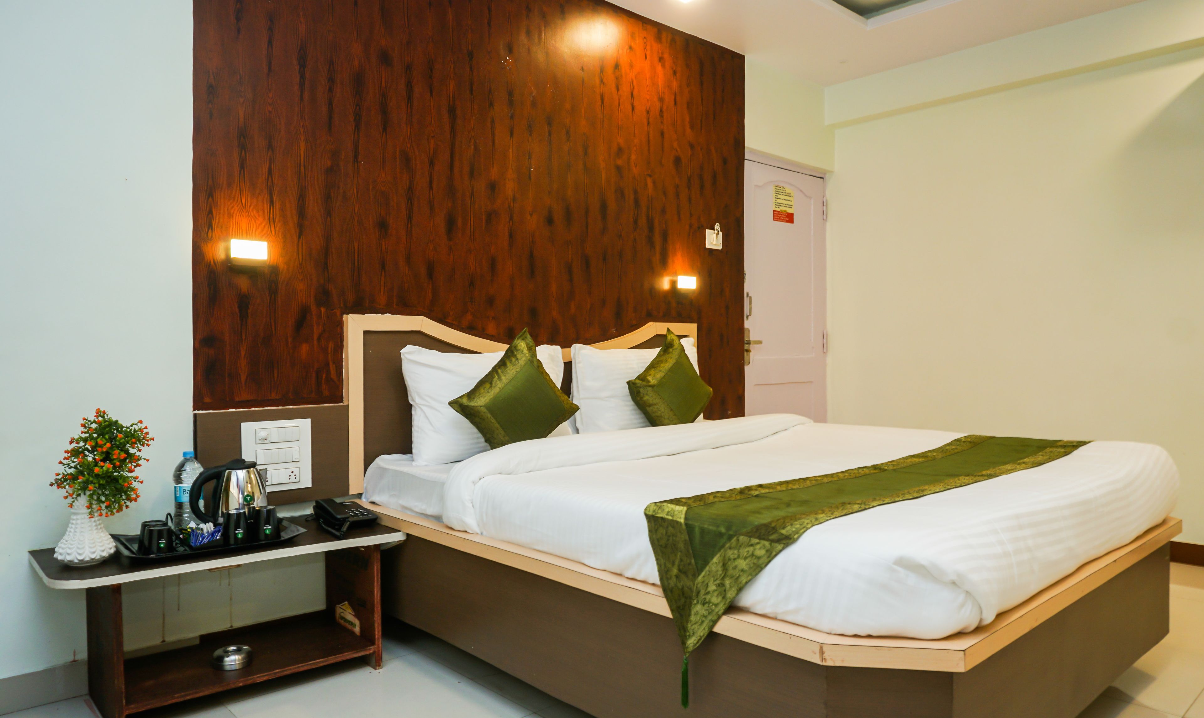 A hotel room with a wooden headboard, a bed with white linens and green pillows, a bedside table with a phone and a vase of flowers, and a nightstand with a lamp and a bottle of water.