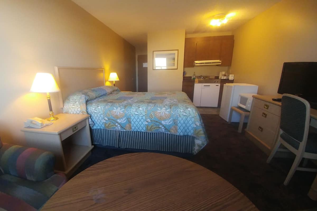A hotel room with a bed, a desk with a lamp, a chair, a small kitchenette, and a television.