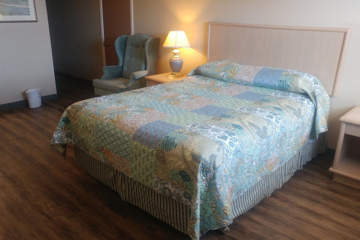 A neatly made bed with a floral quilt, a bedside lamp, a chair, and a small table in a hotel room with wooden flooring.
