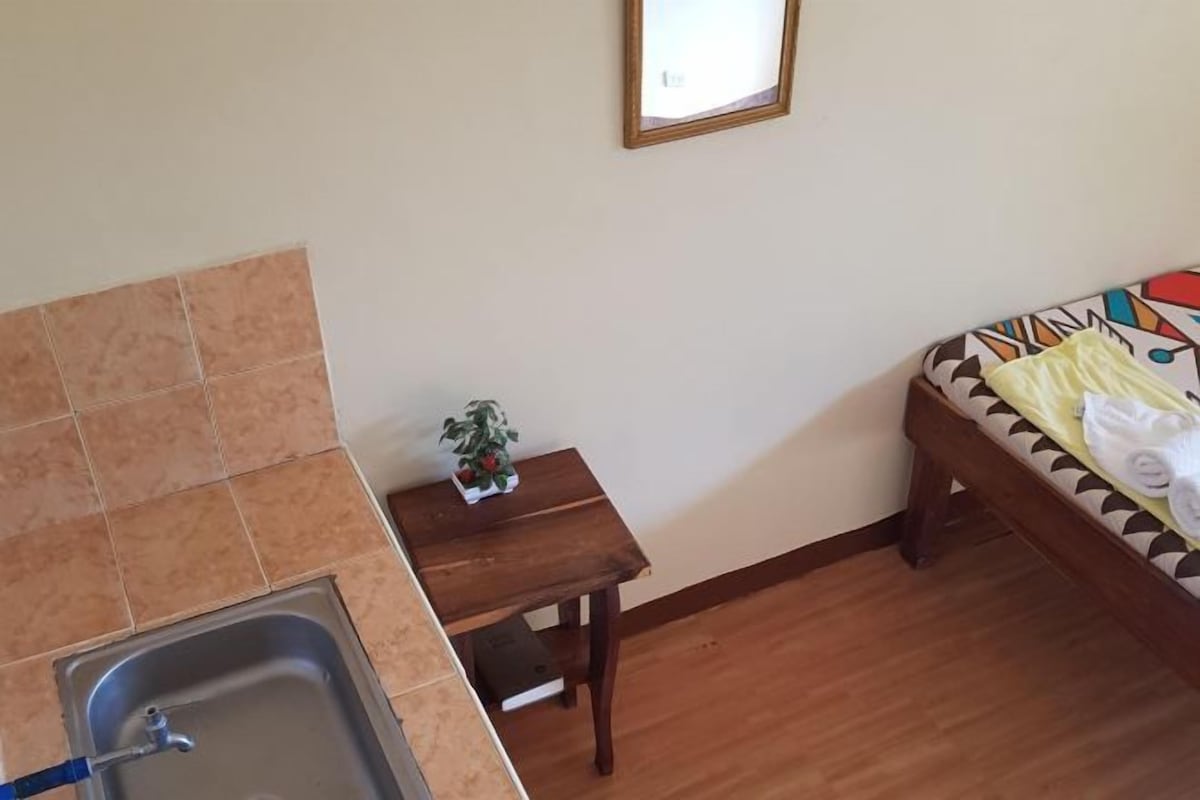 Una cocina pequeña con un lavamanos, una mesa de madera con una planta y una cama con colcha estampada.