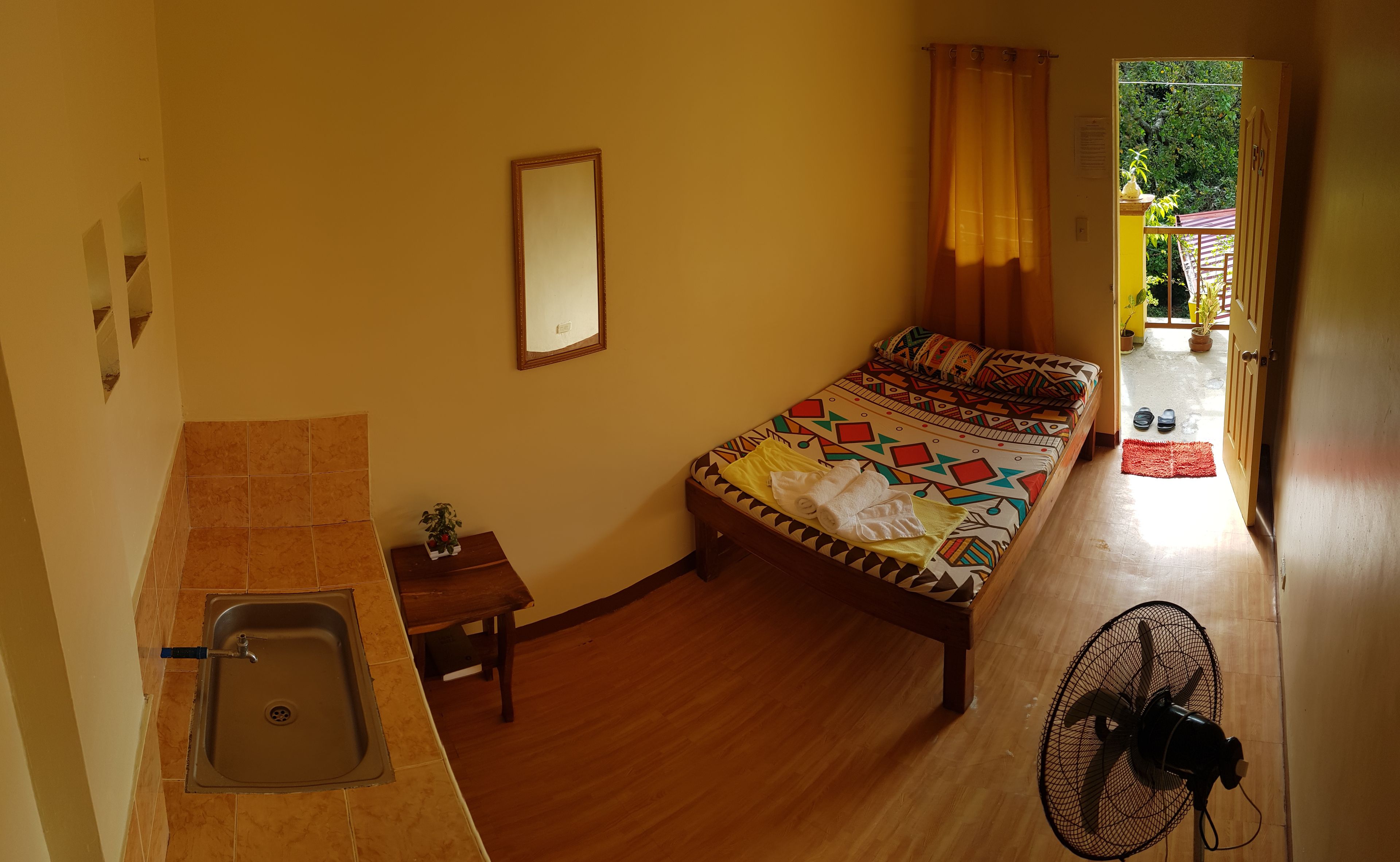 Una habitación con cama, lavamanos, espejo, ventilador y vista al exterior.