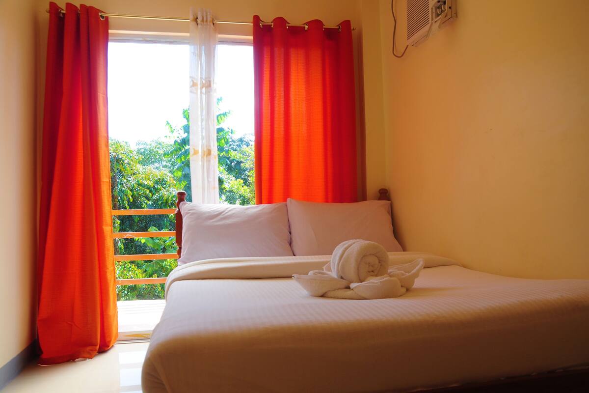A hotel room with a bed, white linens, a window with red curtains, and a view of greenery outside.