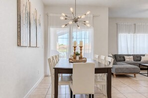 A dining area with a wooden table, white chairs, a chandelier, and a gray sofa in the background.