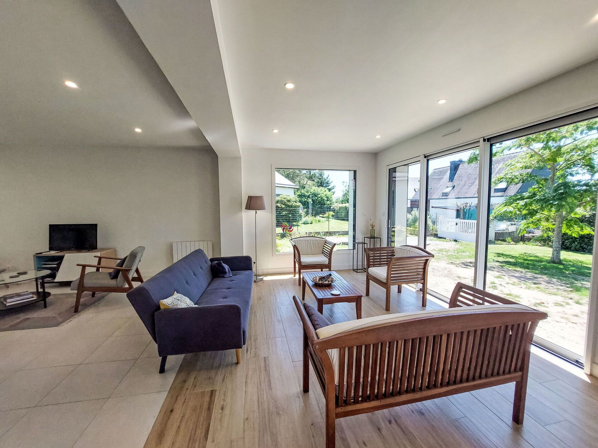Una sala de estar moderna con sofá, sillas de madera, una mesa de centro y televisión. Amplias ventanas ofrecen vistas al exterior.