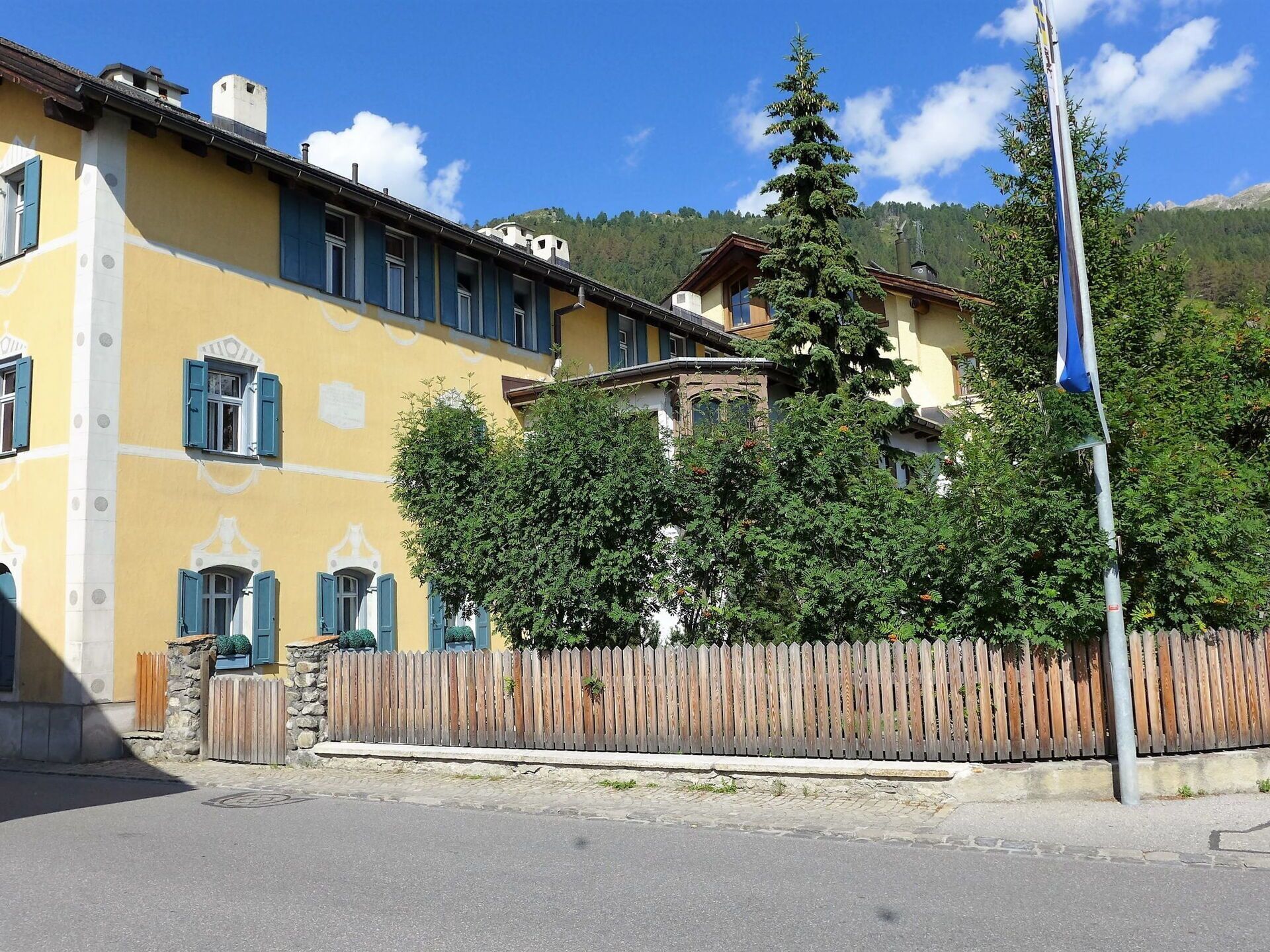 A yellow building with blue shutters, a wooden fence, and a tall tree in front.