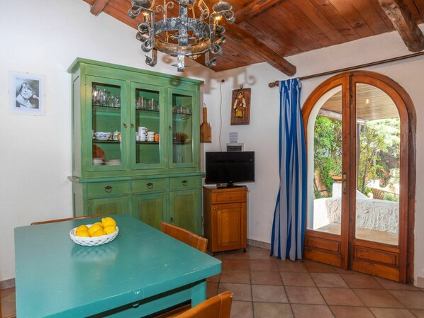 Un comedor con una mesa color turquesa, un mueble con puertas de cristal, un televisor y vistas a un jardín exterior a través de una puerta de madera.