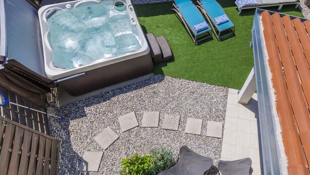A hot tub with a stone wall backdrop, surrounded by a patio with lounge chairs and a small garden area.