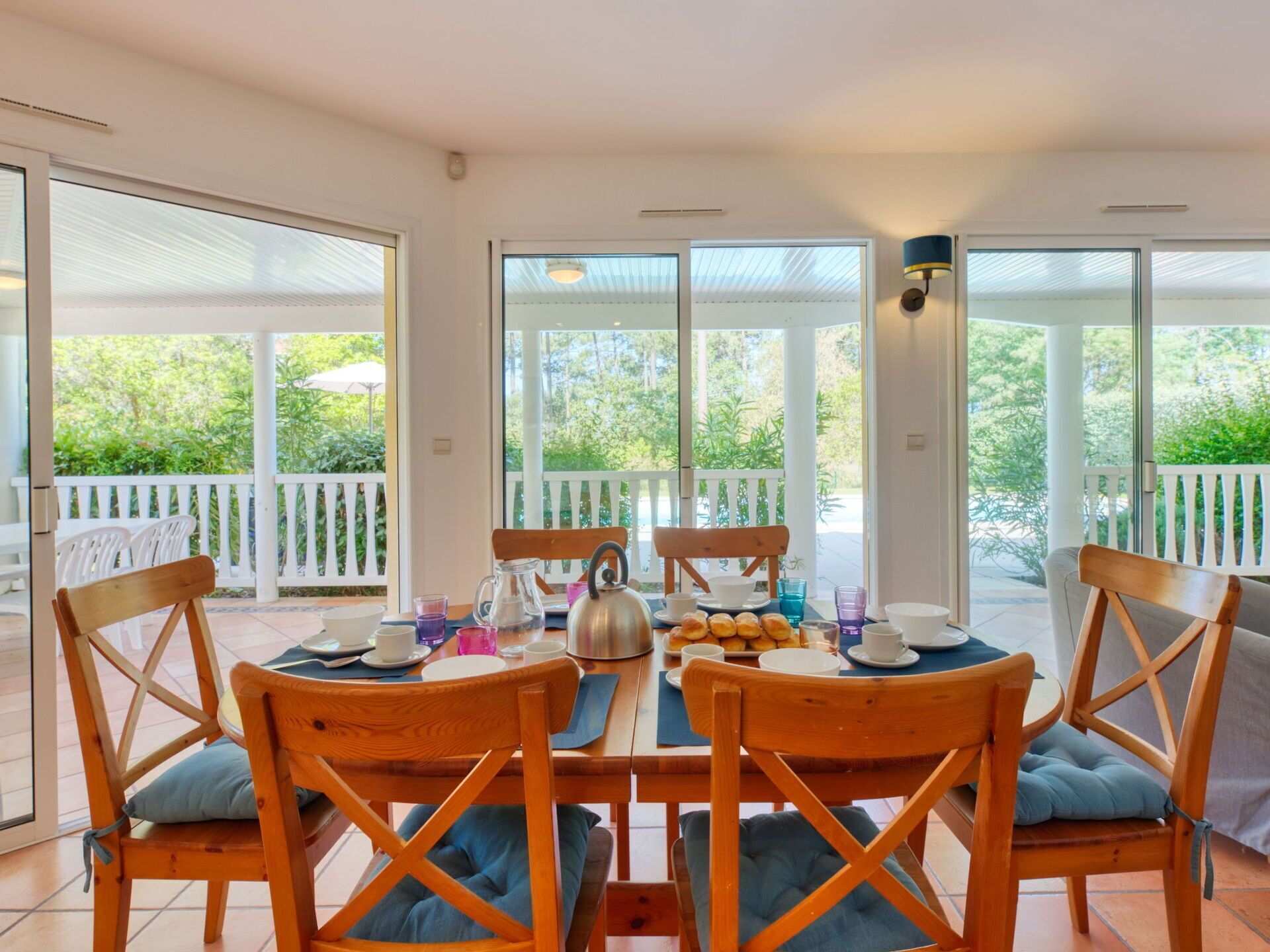 Un comedor con una mesa dispuesta para cuatro personas, rodeada de sillas de madera con cojines azules, ventanas amplias que ofrecen vistas a la vegetación y una baranda blanca en el exterior.