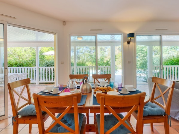 A dining area with a table set for four, surrounded by wooden chairs with blue cushions, large windows offering a view of greenery, and a white railing outside.