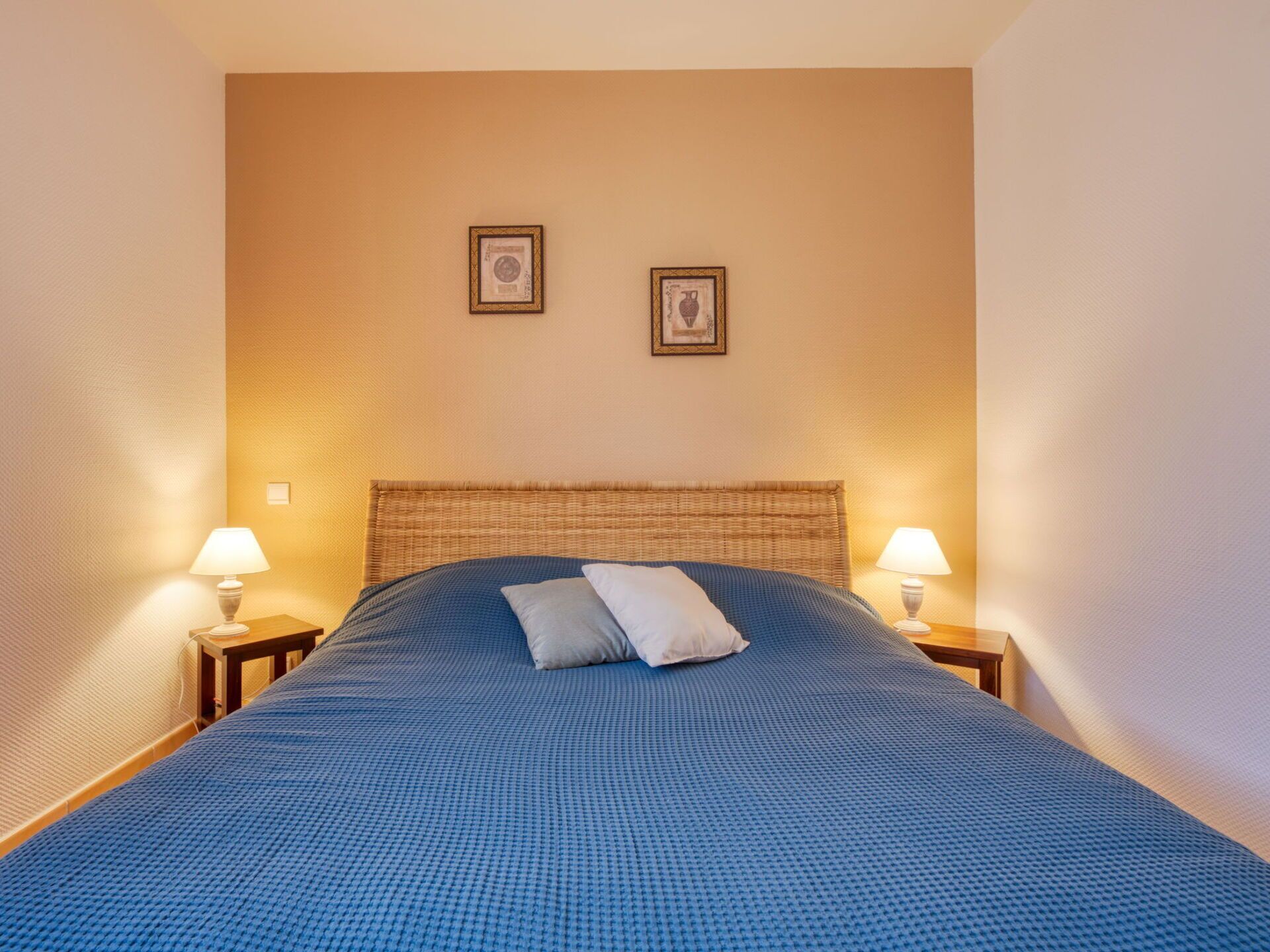 Un lit bien fait, recouvert d’une couverture bleue à motifs, avec deux oreillers et une tête de lit en osier. Deux lampes de chevet sont posées sur des tables de chevet en bois, et deux tableaux encadrés ornent le mur au-dessus du lit.