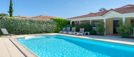 A swimming pool with lounge chairs, a covered patio, and a well-maintained hedge.