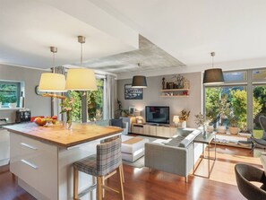 A modern kitchen with a central island, a dining area with a table and chairs, a living room with a sofa and TV, and a large window with a view of greenery.