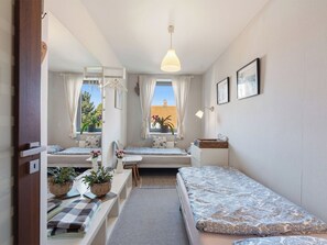 A compact bedroom with a single bed, a small desk, and a chair. There is a window with curtains, a hanging light fixture, and a wall-mounted shelf with a basket.