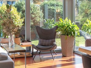Interior moderno com uma janela ampla, uma poltrona estofada cinza, uma cesta de vime com plantas e uma mesa de madeira com objetos decorativos.