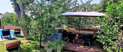 Área de pátio com mesa e cadeiras, cercada por plantas em vasos e uma árvore.