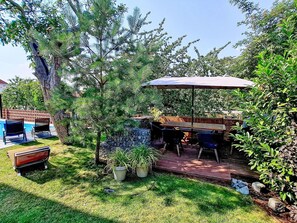 Área de pátio com mesa e cadeiras, cercada por plantas em vasos e uma árvore.
