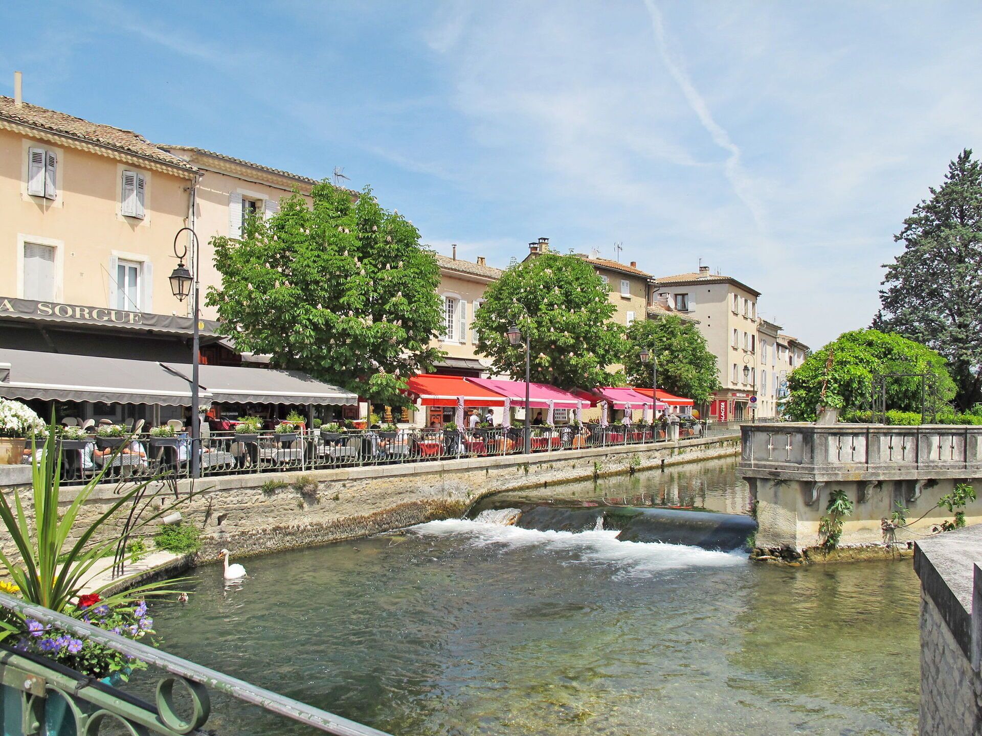 A riverside area with outdoor seating, a bridge, and buildings in the background.