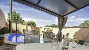 Uma área de estar coberta ao ar livre, com piscina, espreguiçadeiras e uma mesa posta para refeições.