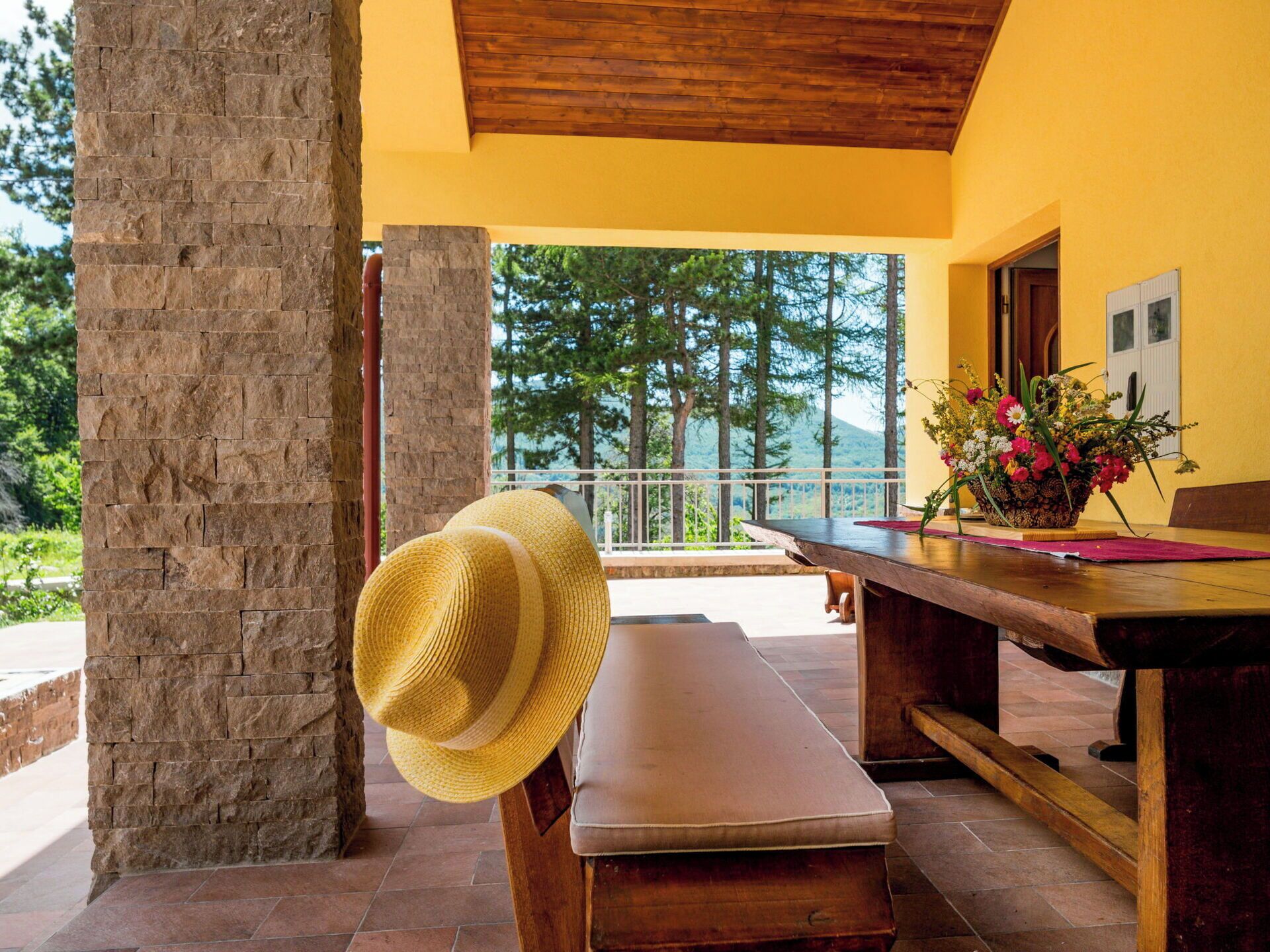 Un banco de madera con un sombrero de paja, una mesa con un florero y vistas a árboles y montañas.
