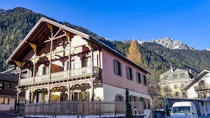 A two-story wooden building with balconies, a stone wall, and a snow-covered ground. - Cosy apartment for 4 people with hot tub, WIFI and TV (Chamonix)