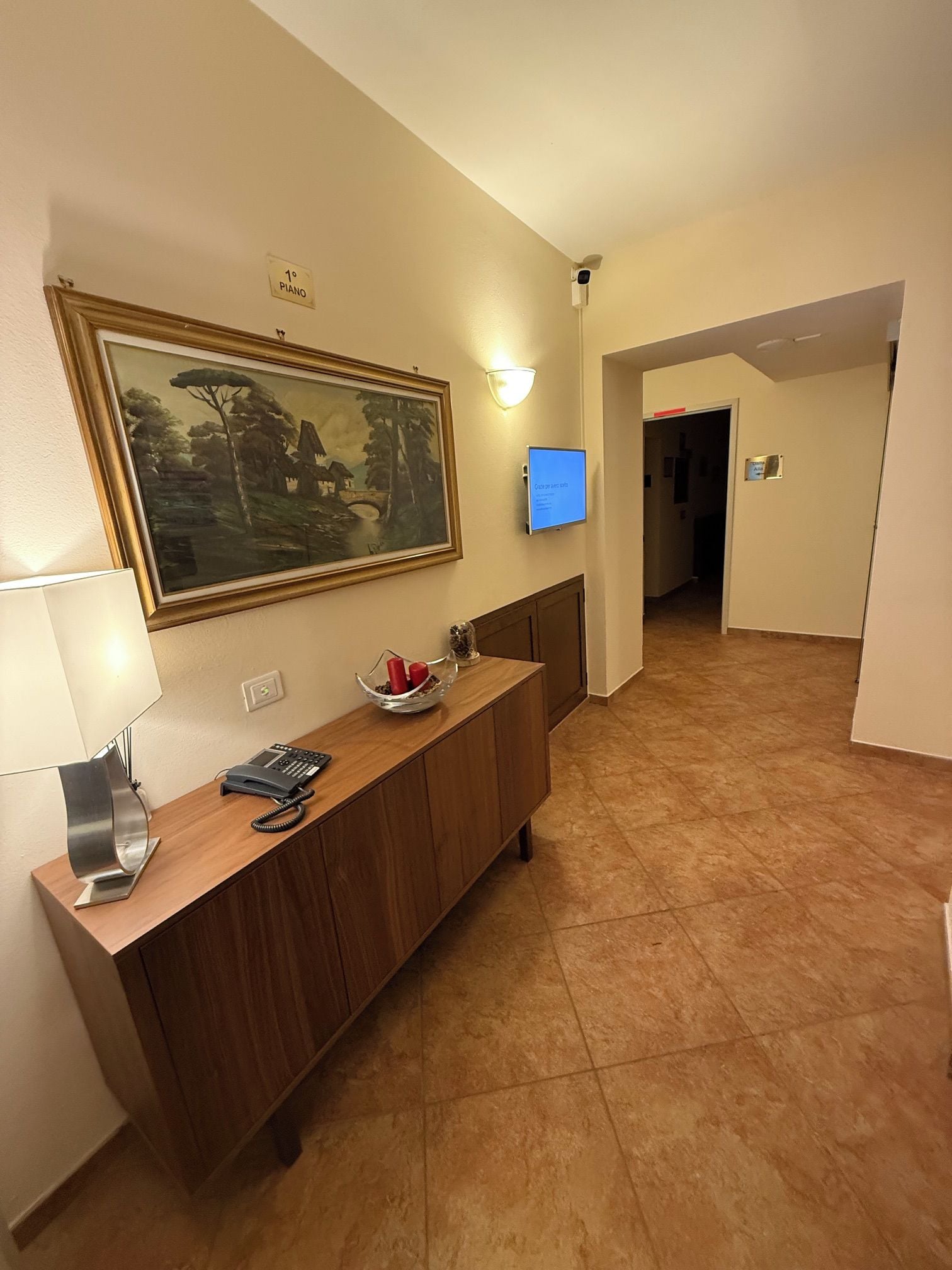 A hotel lobby with a reception desk, a painting, a television, and a sign indicating a piano room.