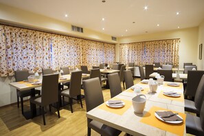 A hotel dining area with multiple tables, chairs, and a large window with curtains. - Hotel Trieste (Tarvisio)