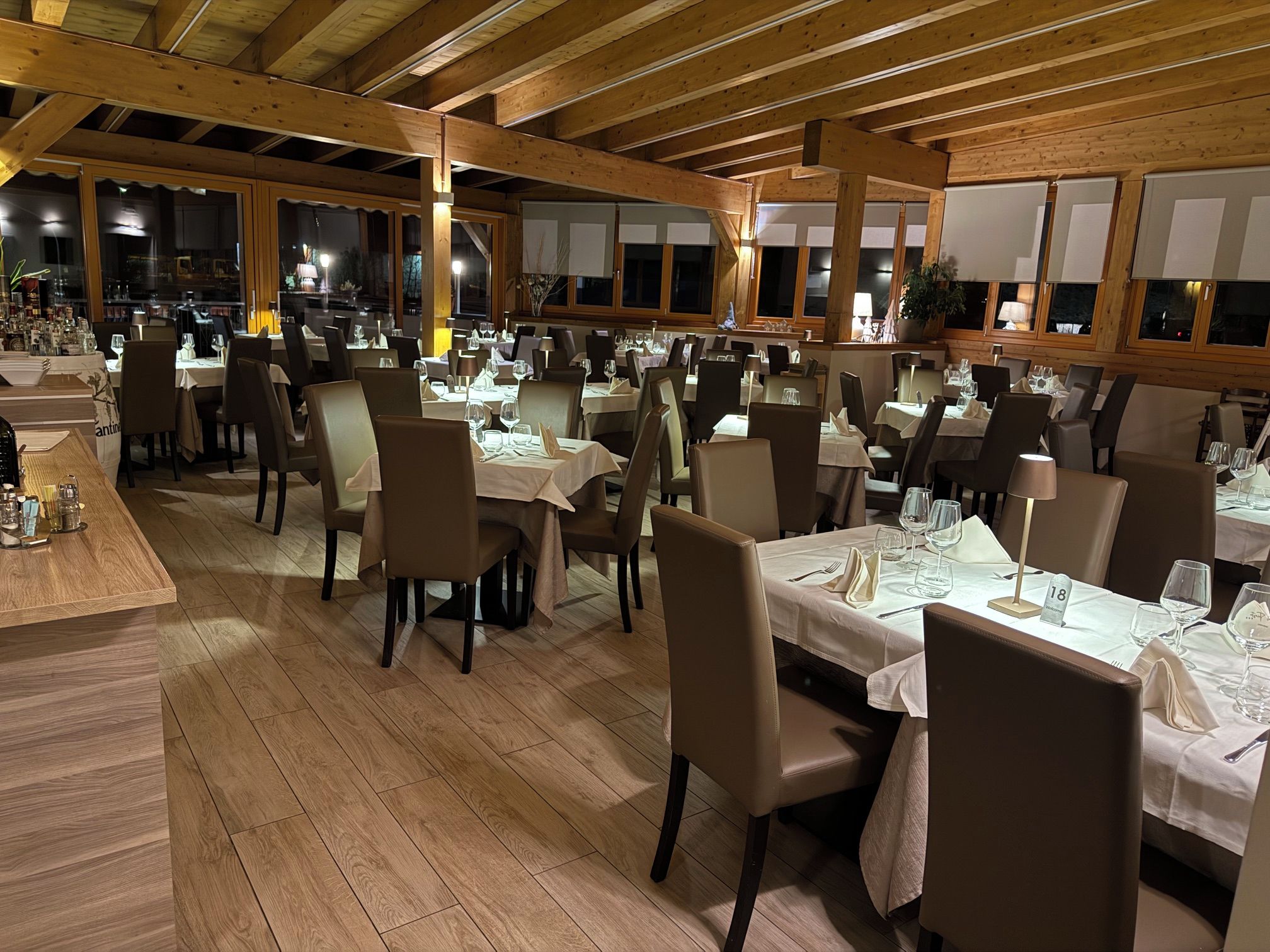 A restaurant with multiple tables and chairs, wooden flooring, and a high ceiling with exposed beams.